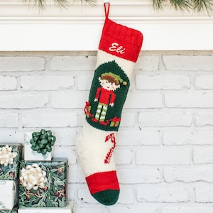 May include: A knit Christmas stocking featuring a red cuff with the name Eli embroidered in white, a central dark green panel with a graphic of an elf, and a red candy cane design on the cream-colored lower section with a dark green toe.