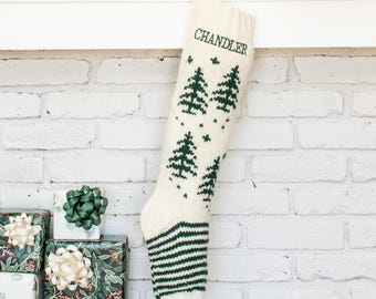 Könnte beinhalten: Ein cremefarbener Strick-Weihnachtsstrumpf mit einem sich wiederholenden Muster aus dunkelgrünen Tannenbäumen und dem Namen CHANDLER, der oben in dunkelgrünem Garn eingestickt ist. Der Zehen- und Fersenbereich zeigt ein dunkelgrün-cremefarbenes Streifenmuster.
