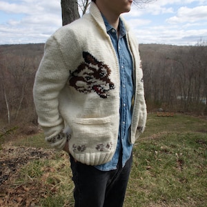 K&ouml;nnte beinhalten: Cremefarbene Strickjacke mit Wolfsmotiv. Der Pullover hat einen Schalkragen, zwei Vordertaschen und eine braune und rote Wolfskopf-Grafik. Die Person tr&auml;gt ein Jeanshemd und eine schwarze Jeans.