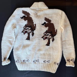 May include: Cream-colored knitted sweater with a collared neckline and long sleeves. The back features two brown wolf designs. The cuffs and hemline are adorned with a repeating floral pattern in brown.
