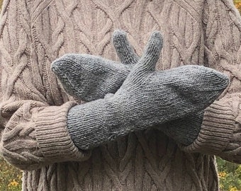 May include: A pair of gray knitted mittens, held in front of a person wearing a brown cable knit sweater.