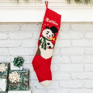 May include: A red knit Christmas stocking featuring a white snowman with a black hat, a green scarf, and a yellow nose. The name Matthew is embroidered in white script at the top. The stocking has a ribbed cuff and a hanging loop at the top edge.