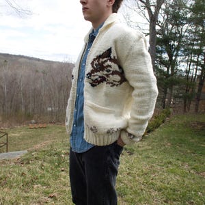 K&ouml;nnte beinhalten: Cremefarbene Strickjacke mit Wolfsmotiv auf der Vorderseite. Der Pullover hat einen Schalkragen, zwei Vordertaschen und einen Knopfverschluss. Die Person tr&auml;gt ein Jeanshemd und eine schwarze Jeans.