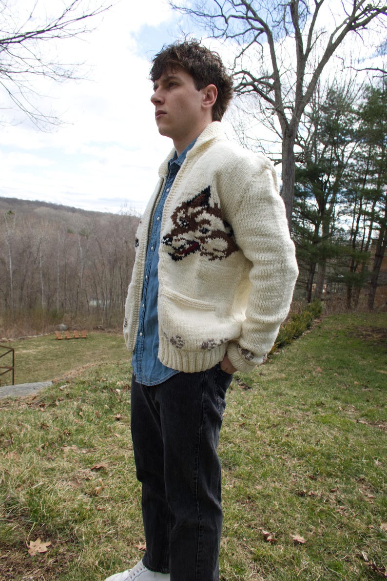 K&ouml;nnte beinhalten: Cremefarbene Strickjacke mit Wolfsmotiv auf der Vorderseite. Der Pullover hat einen Schalkragen, zwei Vordertaschen und einen Knopfverschluss. Die Person tr&auml;gt ein Jeanshemd und eine schwarze Jeans.