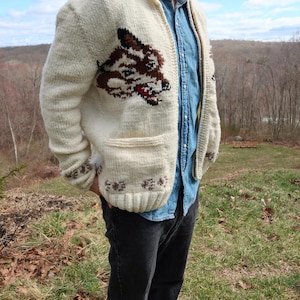 Può includere: Cardigan in maglia grossa color crema con un grande disegno a testa di lupo sul davanti. Il lupo è marrone e nero con accenti rossi. Il maglione ha due tasche anteriori e polsini a coste. La persona indossa una camicia di jeans e jeans neri.