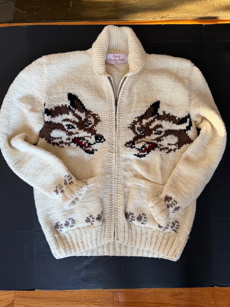 K&ouml;nnte beinhalten: Cremefarbener Rei&szlig;verschluss-Pullover mit zwei Wolfsdesigns auf der Vorderseite. Der Pullover hat einen Schalkragen, zwei Taschen und braune und schwarze Wolfsgesichter. Der Text "Camp Wawby Kella" steht auf dem Kragen.