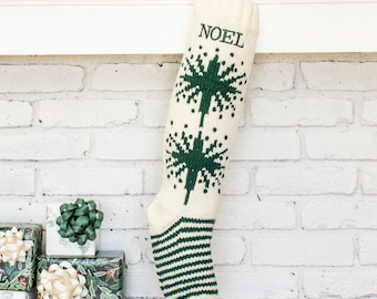 May include: A cream-colored knit Christmas stocking featuring a dark green embroidered starburst pattern and the word NOEL in green text. The foot of the stocking displays a dark green and cream horizontal striped design throughout the lower section.