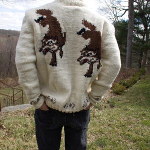 K&ouml;nnte beinhalten: Cremefarbener, handgestrickter Pullover mit einem braunen Cartoon-Charakter-Design auf dem R&uuml;cken. Der Pullover hat einen Rollkragen und gerippte B&uuml;ndchen. Der untere Saum und die Manschetten haben ein dekoratives Muster. Die Person tr&auml;gt dunkle Jeans.
