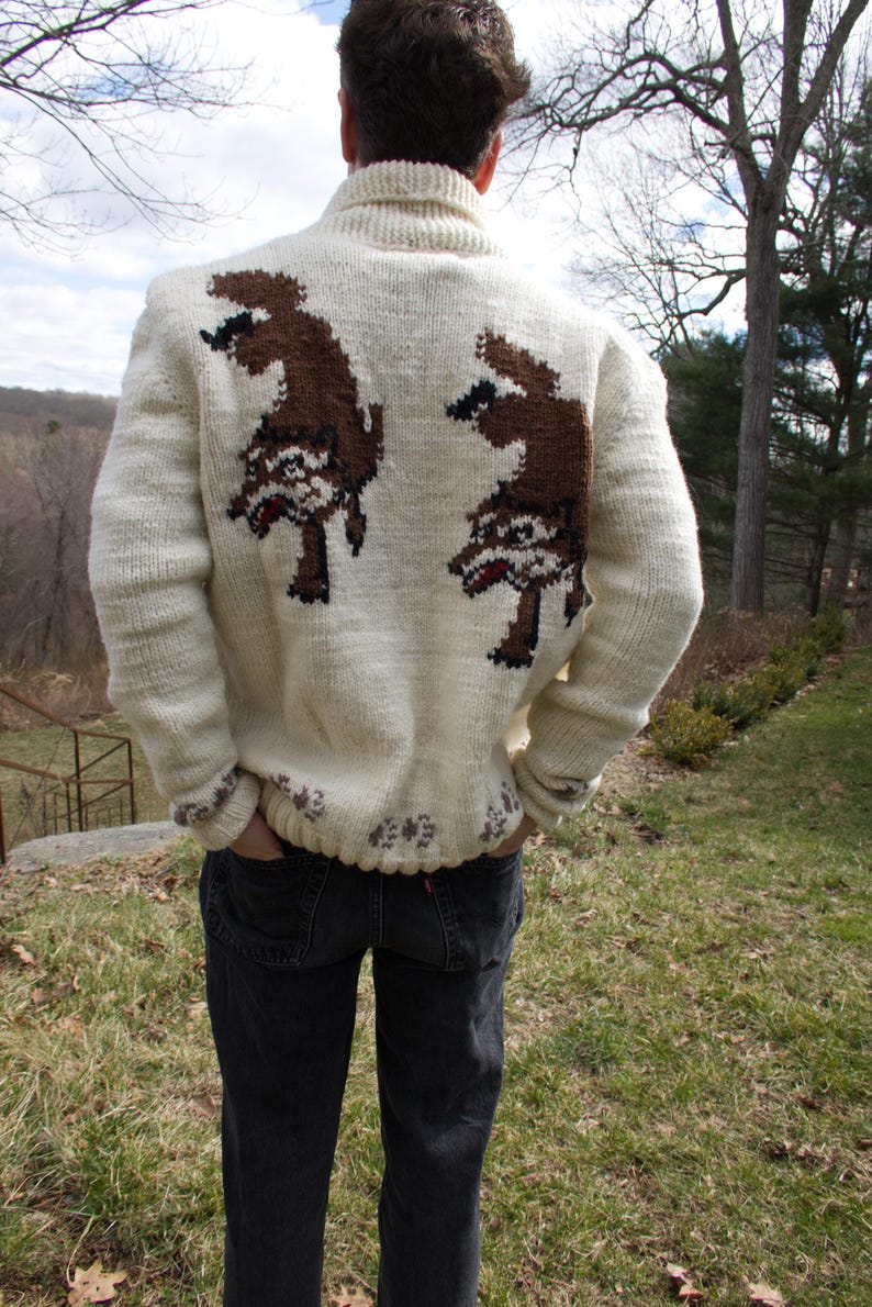 K&ouml;nnte beinhalten: Cremefarbener, handgestrickter Pullover mit einem braunen Cartoon-Charakter-Design auf dem R&uuml;cken. Der Pullover hat einen Rollkragen und gerippte B&uuml;ndchen. Der untere Saum und die Manschetten haben ein dekoratives Muster. Die Person tr&auml;gt dunkle Jeans.