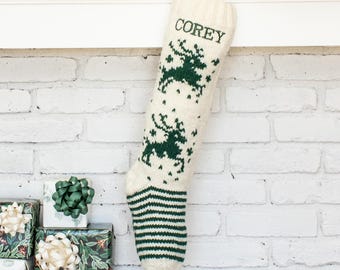May include: A cream-colored knit Christmas stocking featuring a dark green reindeer pattern and the name COREY embroidered in dark green at the top. The toe and heel area of the stocking displays a dark green and cream horizontal striped design.