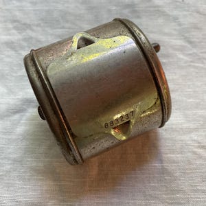 May include: A cylindrical metal object with a hinged lid, showing signs of age and wear. The lid has the number "883237" stamped on it. The metal has a mix of silver and brown tones, giving it a vintage appearance.