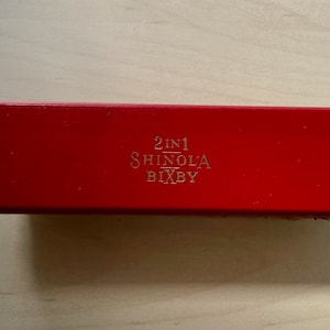 Könnte beinhalten: Eine rote, rechteckige Box mit dem Text "2 in 1 Shinola Bixby" in Gold auf der Oberseite.