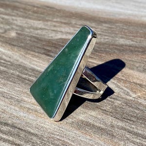 May include: A ring featuring a large, elongated, trapezoidal green stone set in a polished silver bezel. The stone displays natural dark green inclusions and a smooth surface. The silver band is open at the back, showcasing a clean, metallic finish.