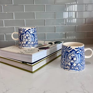 May include: Two ceramic mugs with white handles and a blue floral design. The mugs are placed on a stack of books with a green spine. The background features a light blue tiled wall.