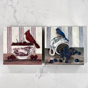 May include: Two square decorative tiles. One features a red cardinal perched above a bowl of cherries. The bowl has a red and white design. The other tile shows a bluebird on a tipped-over teacup spilling blueberries. Both tiles have a striped background.