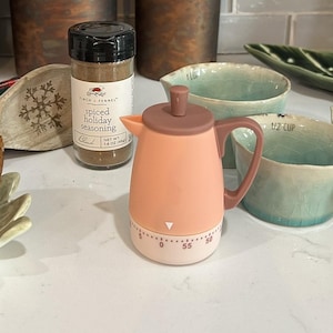 May include: A peach-coloured kitchen timer shaped like a coffee pot with a brown lid and handle. The base features a rotating dial with numbers from 0 to 55. Next to it is a clear spice jar labelled Finch + Fennel spiced holiday seasoning, net wt 45g.