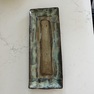 Puede incluir: Una bandeja rectangular de cerámica con un centro texturizado de color marrón tierra y un borde brillante y variado en tonos verde salvia, azul cerceta y marrón oscuro. Los bordes son ligeramente irregulares, mostrando un aspecto artesanal.