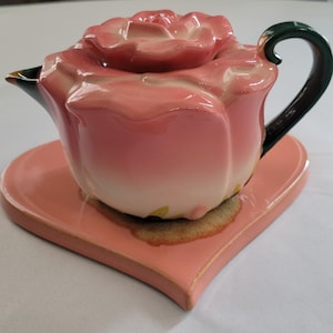 May include: A ceramic teapot shaped like a blooming rose with a matching lid, featuring gradient pink petals that fade to a cream base. It includes a dark green curved handle and a matching heart-shaped saucer in a solid light pink finish.