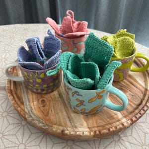 Może przedstawiać: Cztery ceramiczne kubki w kolorach fioletowym, jasnoniebieskim, różowym i żółtym, każdy ozdobiony innym abstrakcyjnym wzorem. Wewnątrz każdego kubka znajduje się złożony, teksturowany, szydełkowany kwadrat z tkaniny w pasującym kolorze: lawendowym, turkusowym, różowym i limonkowym.