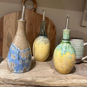 May include: Three ceramic oil bottles with stainless steel spouts. The bottles have unique designs: one with blue and brown glaze, one with yellow and dark blue, and one with yellow and green. They are displayed on a wooden surface.
