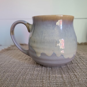 Puede incluir: Una taza de cerámica con cuerpo redondeado y asa curva. El exterior presenta un esmalte reactivo con un degradado que va desde un tono bronceado cálido en el borde hasta un azul grisáceo suave en el centro, con un goteo blanco crema en la base.