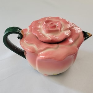 May include: A ceramic teapot shaped like a blooming pink rose. The lid features sculpted rose petals, while the body is rounded with a glossy finish. It includes a dark green curved handle and a small spout with green and brown accents at the tip.