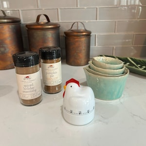 May include: Two clear spice jars with black lids labeled Finch + Fennel Spiced Holiday Seasoning and Pumpkin Pie, a white chicken-shaped kitchen timer with a red comb, and a set of three nested light green ceramic measuring cups with crackle glaze.