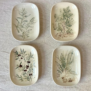 Może przedstawiać: Cztery prostokątne ceramiczne talerze z motywami botanicznymi. Każdy talerz przedstawia inną zielono-brązową ilustrację roślin na kremowym tle. Wzory obejmują paprocie, gałęzie i jagody.