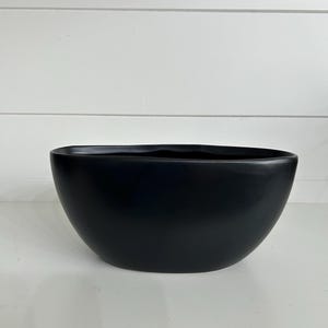 May include: A matte black bowl with a smooth, rounded exterior and a slightly irregular, organic rim. The bowl features a deep, wide basin and a solid, opaque finish, presenting a clean and simple design that is consistent in its dark coloration.