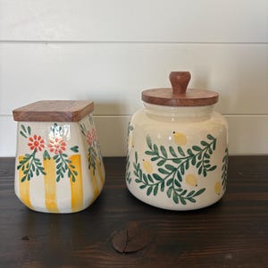 May include: Two ceramic canisters with wooden lids. The left canister is square-shaped with yellow vertical stripes and red floral accents. The right canister is round with a pattern of green leafy branches and small yellow lemon-like shapes.