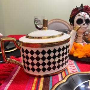 Può includere: Una teiera in ceramica bianca caratterizzata da un motivo a rombi neri attorno al corpo. La teiera include un manico color oro, un coperchio bordato d'oro con pomello dorato e un beccuccio con punta dorata. Il design è pulito, geometrico e simmetrico.