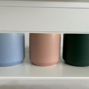 May include: Three cylindrical plant pots with a smooth matte finish, arranged in a row. The pot on the left is light blue, the center pot is soft pink, and the pot on the right is a deep forest green. Each container has a simple, clean design.