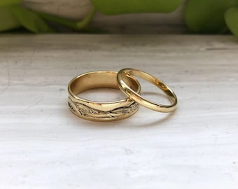 Puede incluir: Dos alianzas de oro, una con un diseño texturizado y la otra lisa, sobre una superficie de madera blanca.