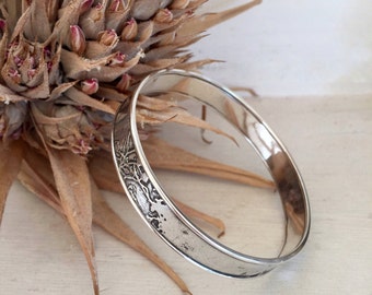 May include: A silver-colored bangle bracelet with an engraved design. The bracelet is round and has a textured surface. The background includes dried, brown plant matter.