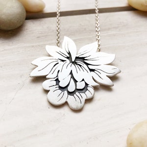 May include: A silver-colored floral pendant necklace. The pendant features a layered flower design with detailed petal outlines. The necklace chain is also silver and delicate, creating an elegant accessory.
