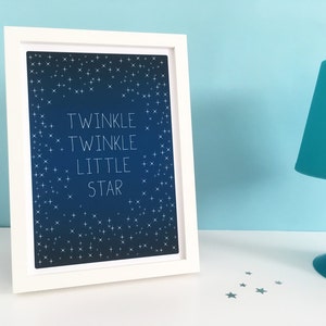 May include: A framed print with the words "Twinkle Twinkle Little Star" in white on a dark blue background, surrounded by white star illustrations. The white frame sits on a white surface next to a teal lamp.