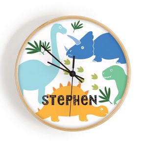 May include: A circular wall clock with a light wood frame. The clock face displays a white background with cartoon dinosaurs in blue, green, and orange. The name "STEPHEN" is printed in black letters on the clock face.