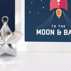 May include: A silver star-shaped ornament with a hanging loop is positioned beside a framed print. The print shows a red rocket with a yellow flame against a dark blue backdrop, with the words "TO THE MOON & BACK."