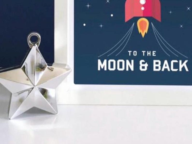 May include: A silver star-shaped ornament with a hanging loop is positioned beside a framed print. The print shows a red rocket with a yellow flame against a dark blue backdrop, with the words "TO THE MOON & BACK."