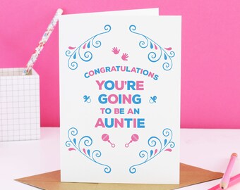 May include: A white card with the message "Congratulations You're Going To Be An Auntie" in pink and blue. The card features blue and pink decorative swirls, baby rattles, and handprints. It rests on a brown envelope.