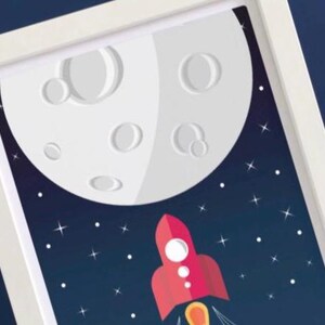 May include: Framed art print depicting a red rocket ship ascending towards a large, white moon with craters. The background is a dark blue space with white stars. The artwork is framed in a white border.