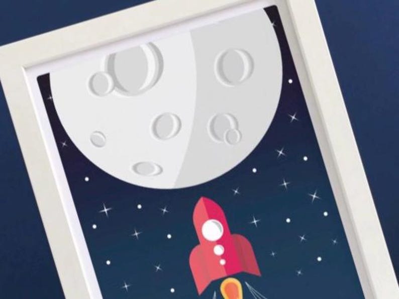 May include: Framed art print depicting a red rocket ship ascending towards a large, white moon with craters. The background is a dark blue space with white stars. The artwork is framed in a white border.