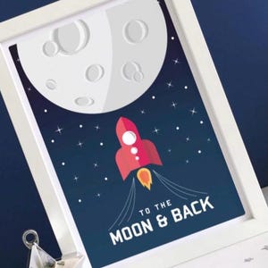 May include: Framed art print showing a red rocket taking off towards a large, white moon. The background is dark blue with stars. Text at the bottom reads "TO THE MOON & BACK". The print is in a white frame.