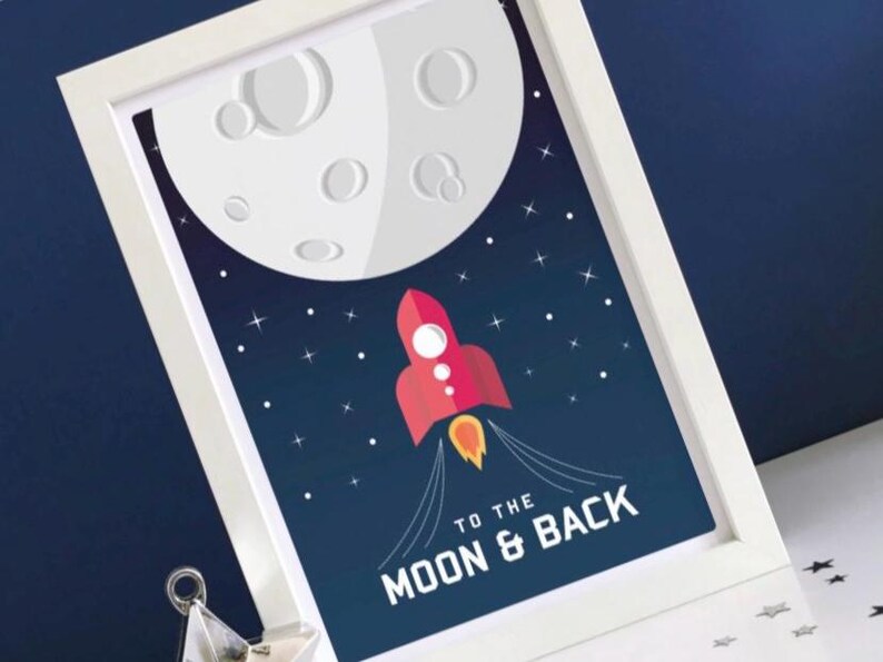 May include: Framed art print showing a red rocket taking off towards a large, white moon. The background is dark blue with stars. Text at the bottom reads "TO THE MOON & BACK". The print is in a white frame.