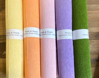 Könnte beinhalten: Fünf Rollen deutsches Floristenkrepppapier in Gelb, Pfirsich, Rosa, Lila und Grün. Jede Rolle hat ein weißes Etikett mit der Aufschrift "Petals & Pearls" und "German Florist Crepe". Das Papier hat eine strukturierte, gekreppte Oberfläche.