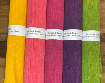 Könnte beinhalten: Fünf Rollen farbiges Krepppapier in Gelb, Rosa, Magenta, Lila und Grün. Jede Rolle ist mit "Petals & Pearls German Florist Crepe" beschriftet. Das Papier hat eine strukturierte, gekreppte Optik, ideal zum Basteln und für Blumenarrangements.