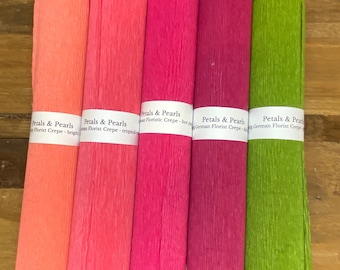 Könnte beinhalten: Fünf Rollen farbenfrohes Floristenkrepppapier in Korallen-, Rosa-, Fuchsia-, Magenta- und Grüntönen. Jede Rolle ist mit dem Text "Petals & Pearls" beschriftet. Die Rollen sind auf einer Holzoberfläche angeordnet.