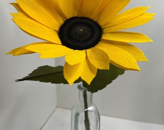 May include: A handcrafted paper sunflower with bright yellow petals and a dark brown center. The flower has green leaves and is displayed in a clear glass bottle. The sunflower is a decorative item.
