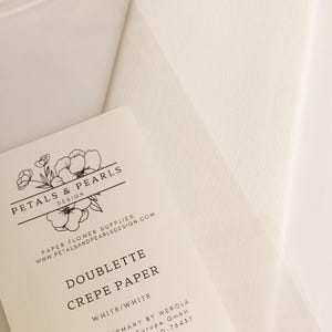 May include: A package of white Doublette crepe paper measuring 9.8 inches by 98 inches. The label features the text Petals & Pearls Design, Paper Flower Supplies, and manufacturer details including the address in Germany for Werola Seaman Paper.