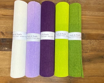 May include: Five rolls of florist crepe paper in various colors: antique white, pastel lilac, dark purple, may green, and green. Each roll has a label that reads "Petals & Pearls Florist Crepe". The crepe paper is textured and rolled up.